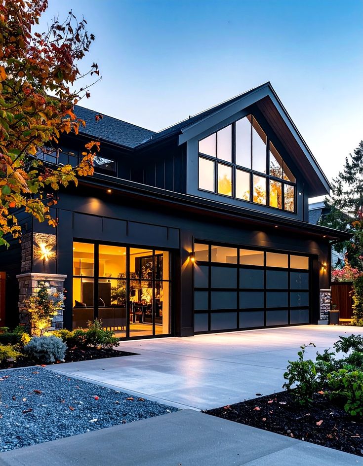 Modern two-story house with large windows and warm interior lighting at dusk, surrounded by autumn foliage.