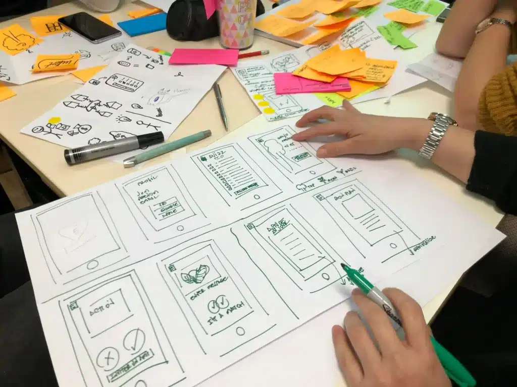 Hands sketching app wireframes with colorful sticky notes and design sketches on a desk.