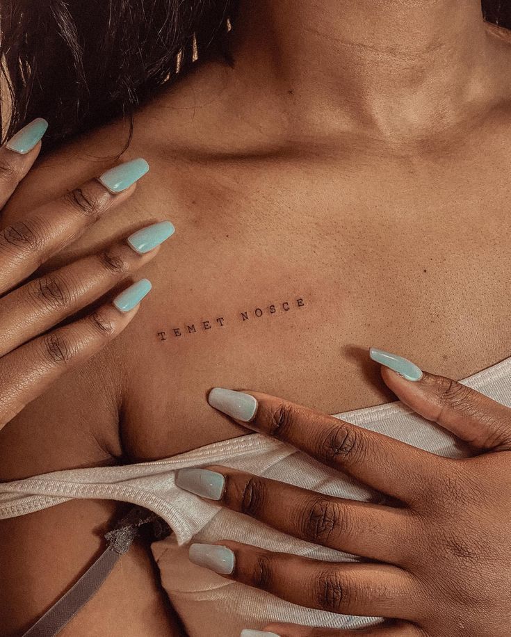 Close-up of a collarbone tattoo reading Temet Nosce, with hands showing pastel blue nails for a stylish look.