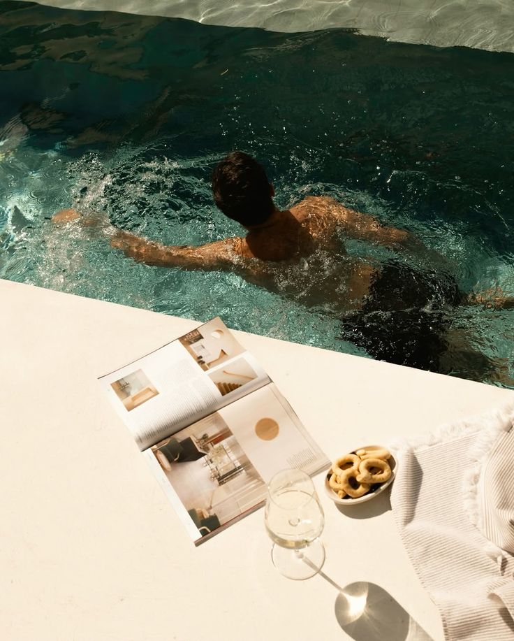 Man swimming in a pool beside a lounging area with a magazine, snacks, and a glass of wine on a sunny day.