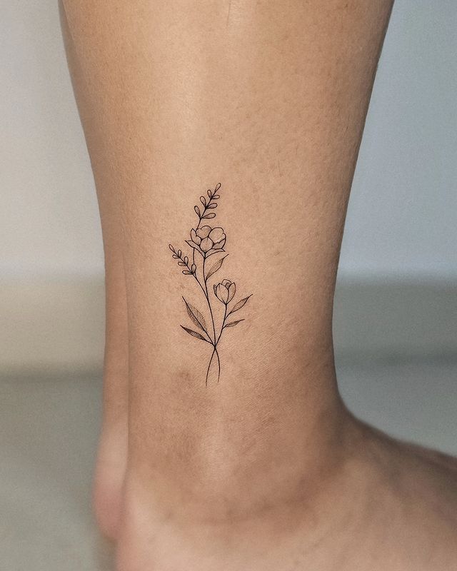 Delicate floral ankle tattoo with fine black linework, showcasing small blossoms and leaves on skin.