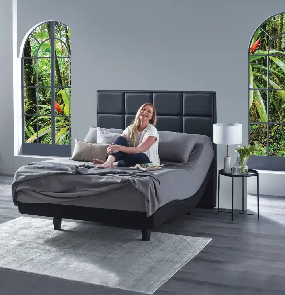 Woman relaxing on a modern adjustable bed in a bright room with a lush garden view through arched windows.