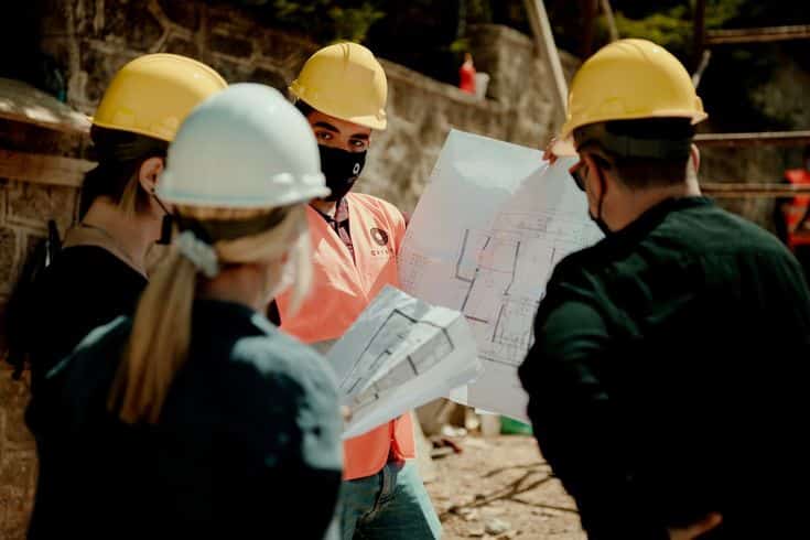 Construction team reviewing blueprints at a building site, wearing protective helmets and masks.