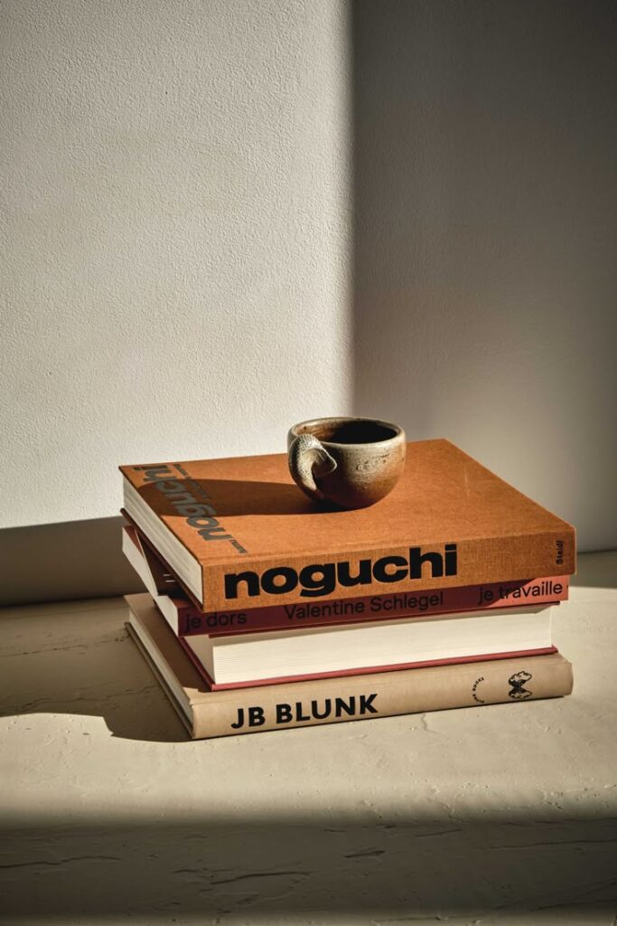 Stack of art books with a ceramic cup on top, placed on a sunlit table against a minimalist background.