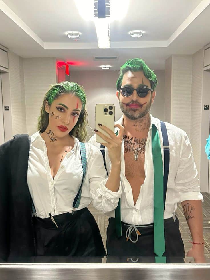 Couple dressed as iconic characters with green hair and makeup taking a mirror selfie in a hallway.
