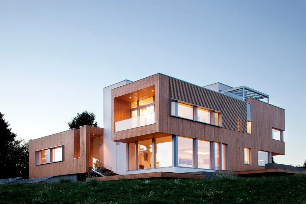 Karuna-House Modern two-story house with large windows, wooden panels, and evening glow, set against a clear sky backdrop. | Sky Rye Design Modern two-story house with large windows, wooden panels, and evening glow, set against a clear sky backdrop.
