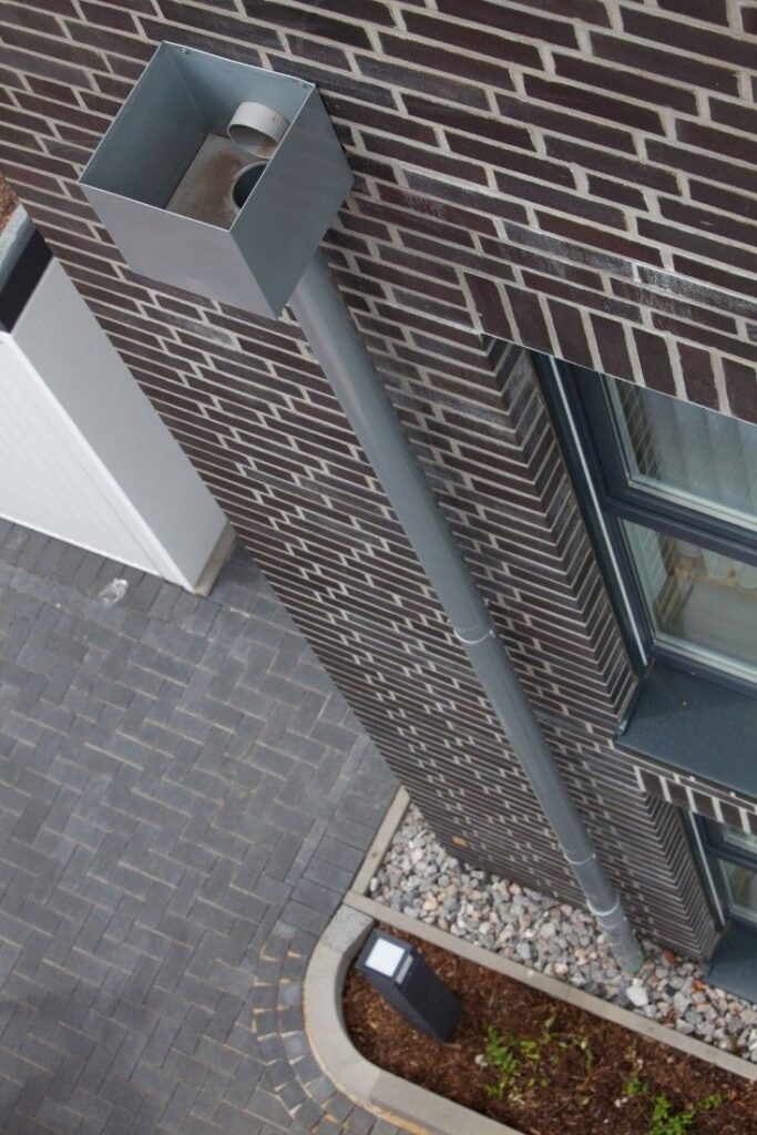 Modern downspout for rainwater drainage on a brick building exterior with surrounding landscape.