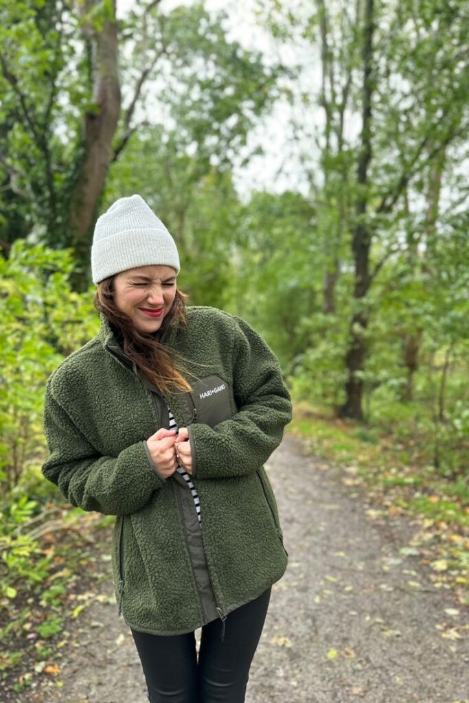Woman in green jacket and white beanie smiling on a wooded path.