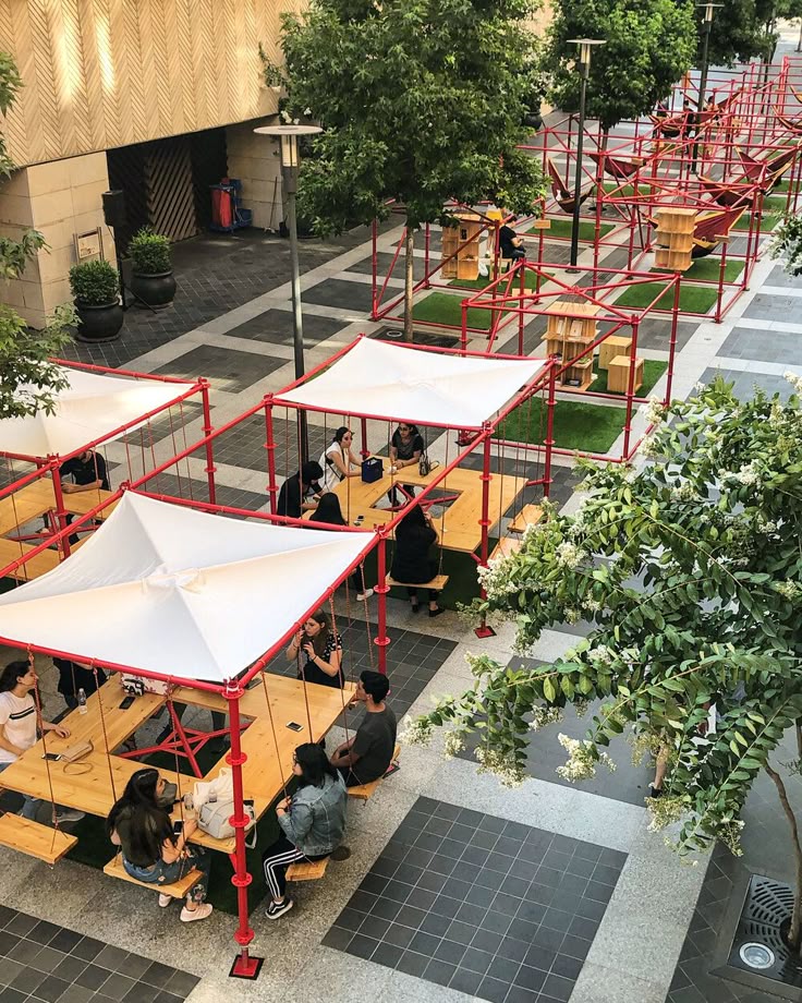 People socializing at outdoor picnic tables under red structures with white canopies in an urban setting.