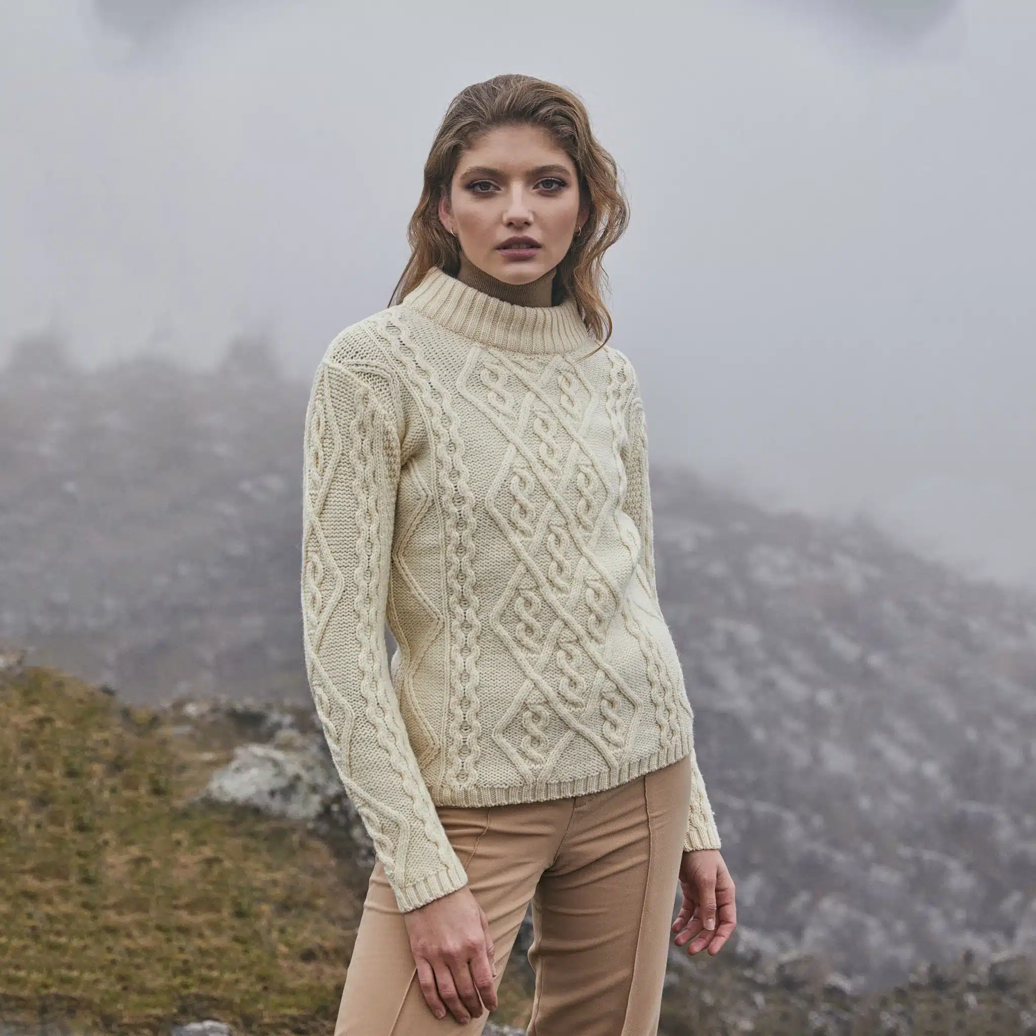 ML905-100-Ladies-Aran-Cable-Knit-Sweater-Lifestyle-1-SAOL_1024x10242x-1 Woman in a cream knit sweater and beige pants stands in misty mountain landscape. | Sky Rye Design Woman in a cream knit sweater and beige pants stands in misty mountain landscape.
