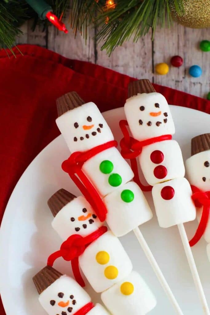 Festive marshmallow snowman pops with candy and chocolate decorations on a plate, adorned with holiday patterns.