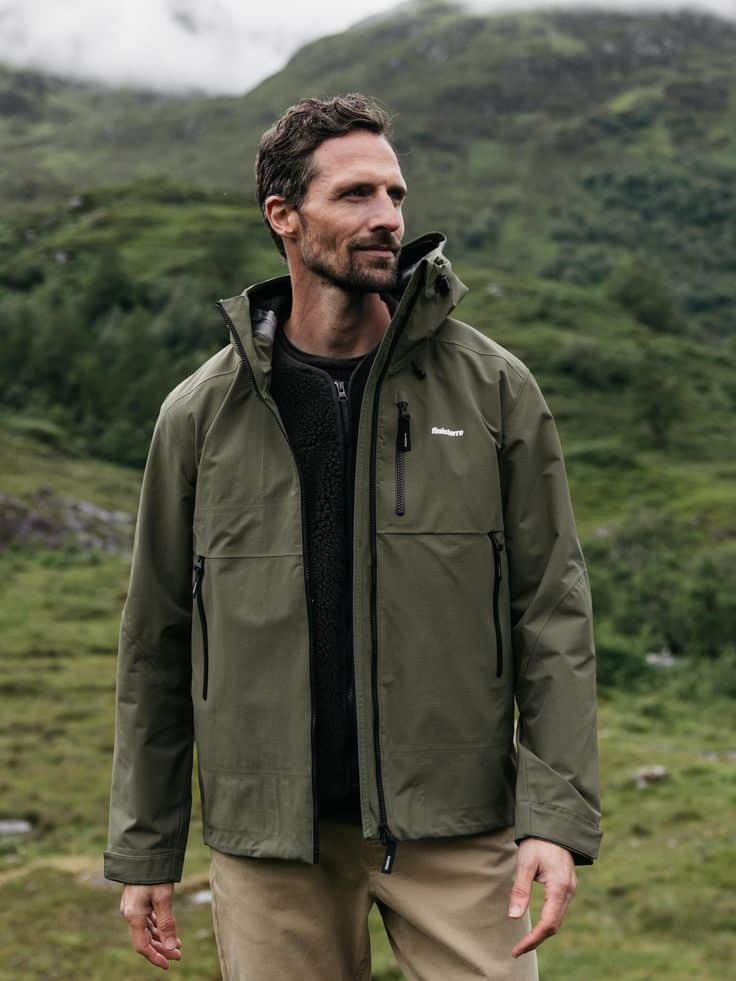 Man in green jacket standing outdoors with a scenic mountain background.