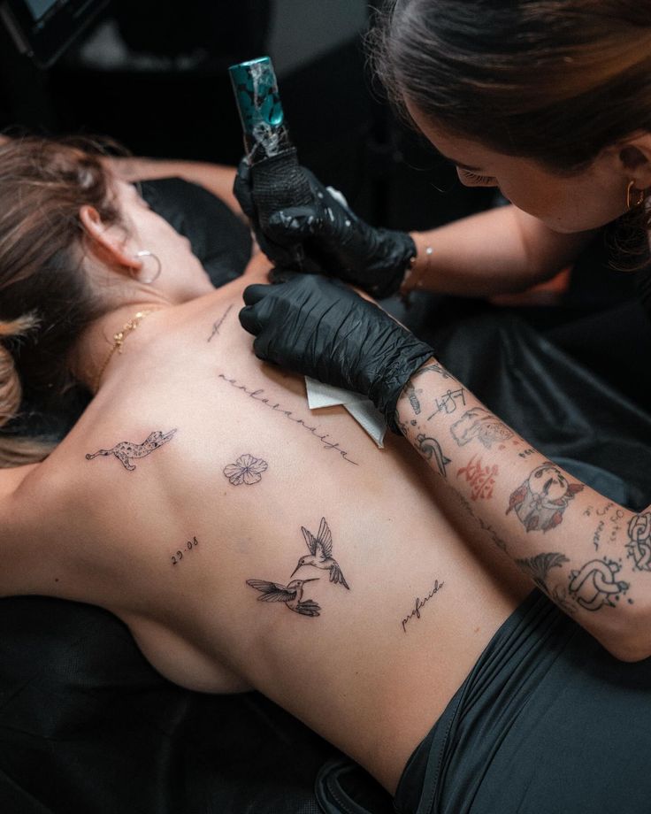 Tattoo artist creating detailed designs on a woman's back, featuring birds and script.