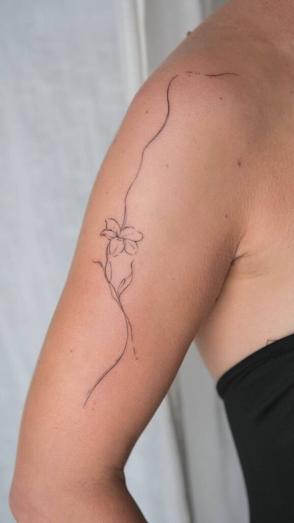 Delicate floral line tattoo on a person's upper arm, featuring a single flower and flowing lines.