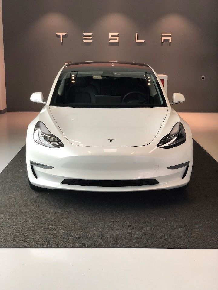 Front view of a white Tesla car in a showroom, emphasizing its sleek design and the Tesla logo on the wall.