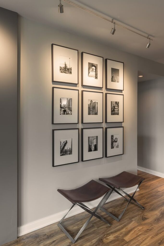 Wall with nine framed black and white photographs and two brown stools on wooden floor under track lighting.