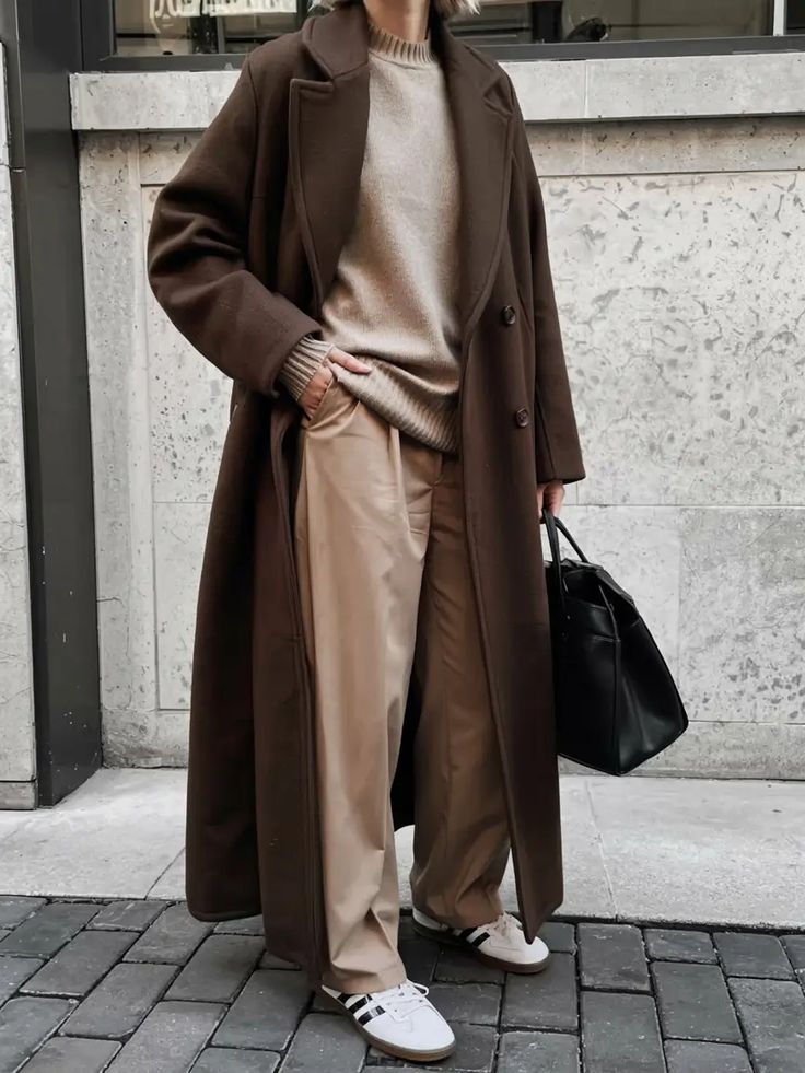 Trendy woman in a long brown coat, beige sweater, and pants, holding a black bag and wearing white sneakers.