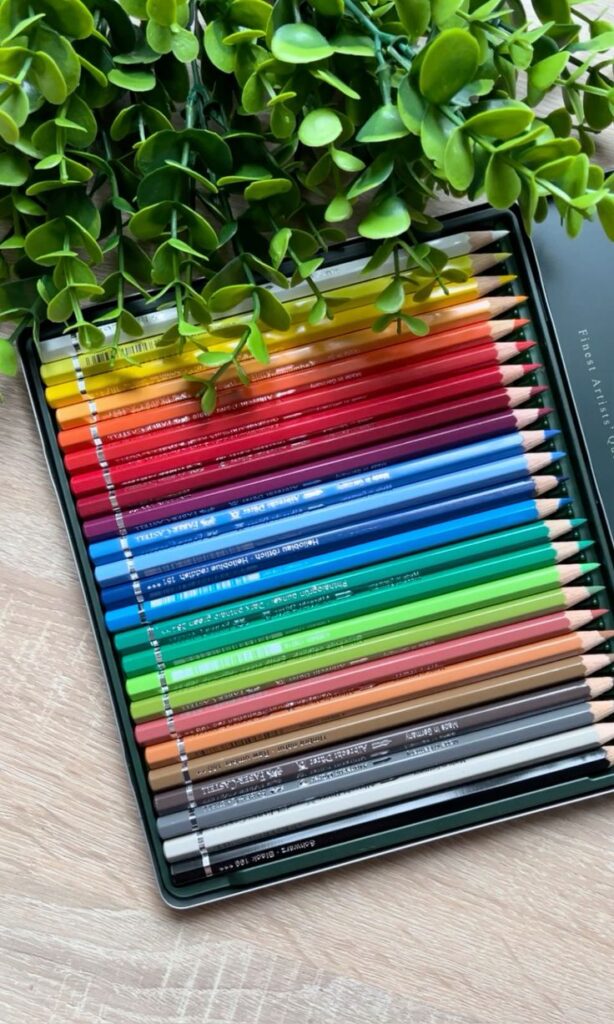 Assorted colored pencils in a black tin, arranged beside green leaves on a wooden surface.