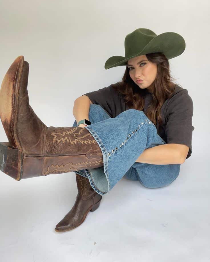 New cowboy boots on the website for all my lil cowgirls out there the real deal or in spirit_ Get them while theyre hot 🤠🔥 1 Woman in cowboy hat and boots sitting cross-legged, wearing jeans and a dark shirt, making a pouty expression. | Sky Rye Design Woman in cowboy hat and boots sitting cross-legged, wearing jeans and a dark shirt, making a pouty expression.