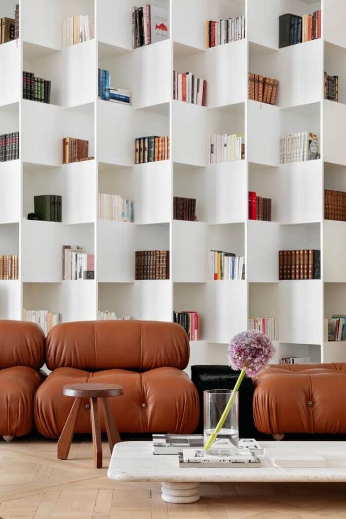 Modern living room with brown leather sofas, white bookshelf filled with books, and a flower vase on a stone table.
