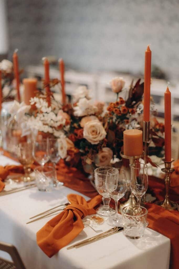 Elegant autumn-themed table setting with orange candles, floral centerpiece, and rust napkins, perfect for a cozy dinner.
