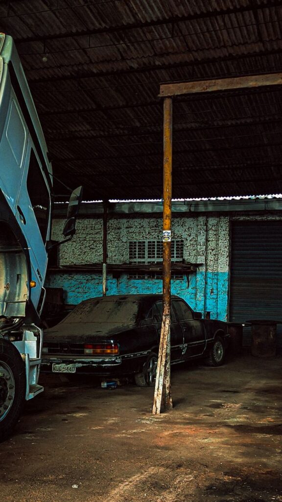 A dusty car and a truck parked in a dimly lit, weathered garage with textured walls and a corrugated roof.