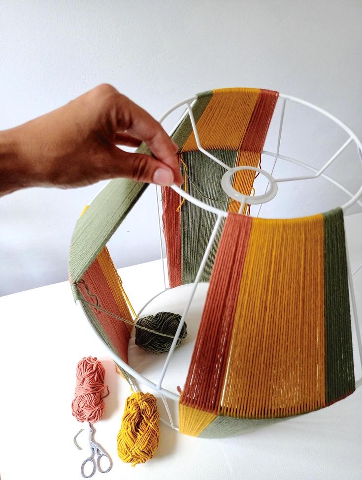 Hand crafting a colorful yarn lampshade on a wire frame with scissors and yarn balls nearby.