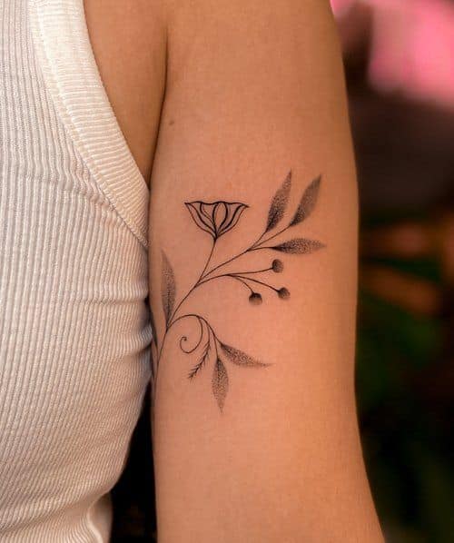 Minimalist floral tattoo on upper arm, depicting delicate leaves and buds in black ink, worn by person in white top.
