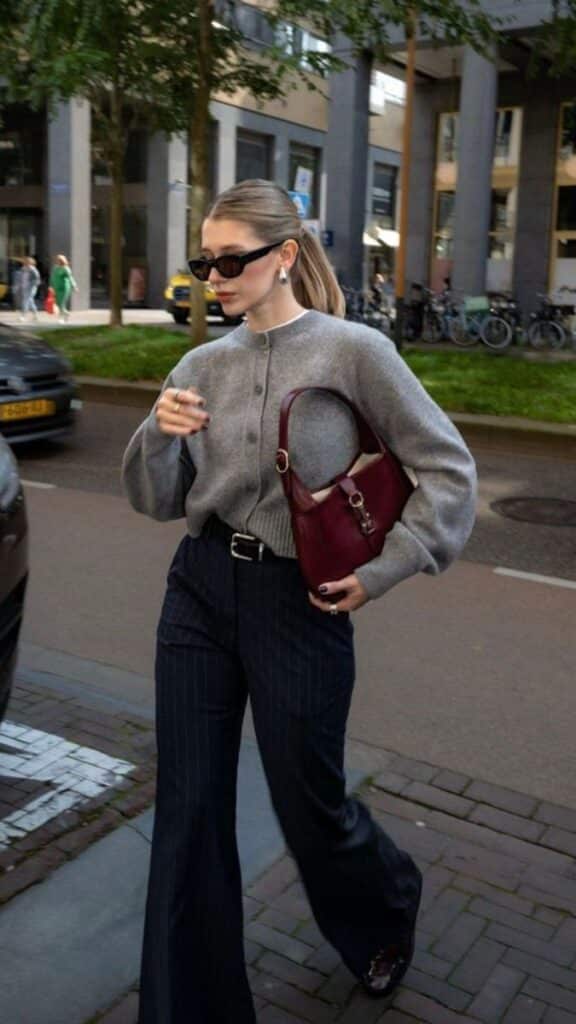 Woman in stylish gray sweater and sunglasses carries maroon purse, walking on city street.