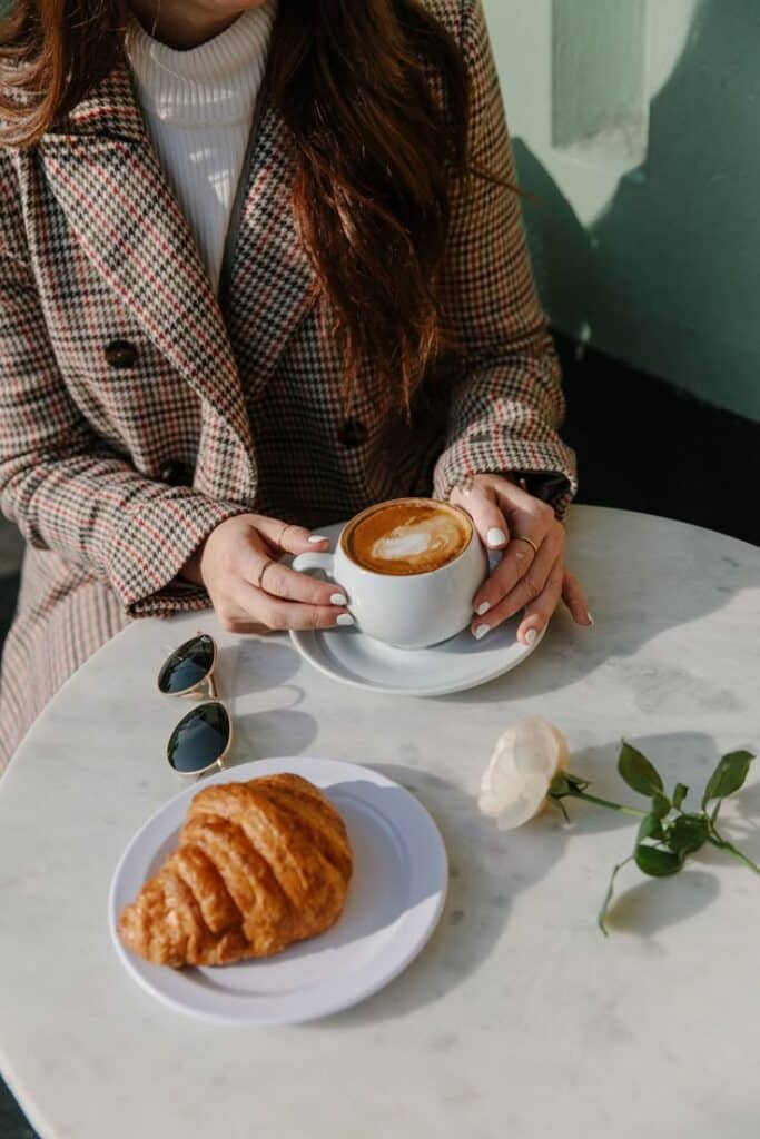 Woman in plaid coat enjoys coffee with a croissant at a marble table, sunglasses and rose nearby.