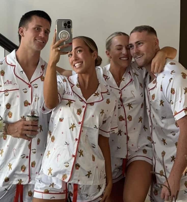 Group photo of four friends in matching holiday-themed pajamas, smiling and taking a selfie indoors.