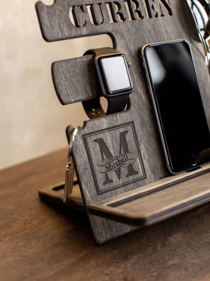 Wooden organizer stand holding a smartwatch, smartphone, and keys, engraved with initials, on a wooden surface.