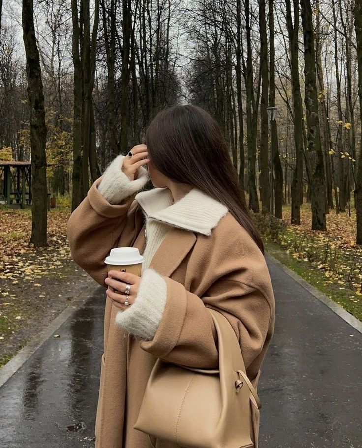 Woman in a tan coat holding coffee, walking through a tree-lined path on a crisp autumn day.