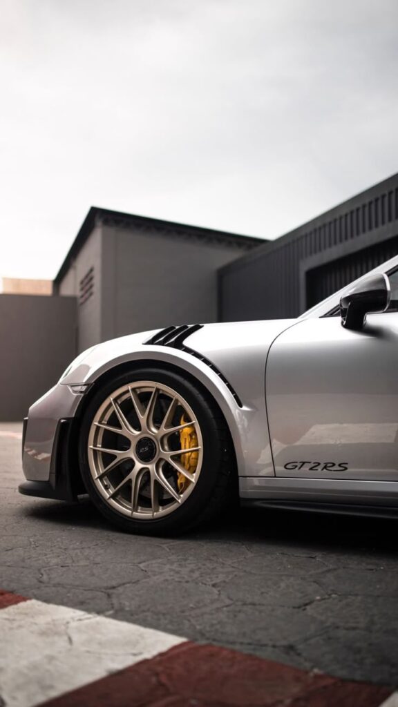 Silver GT2 RS with yellow brake calipers, parked in front of a modern building, showcasing sleek wheel design.