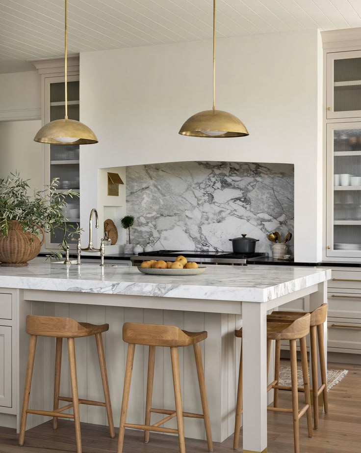 Modern kitchen with marble island, wooden stools, gold pendant lights, and elegant backsplash.