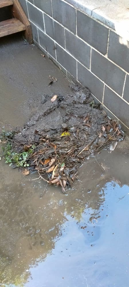 Mud and debris pile against brick wall corner after rainfall, illustrating drainage issue.