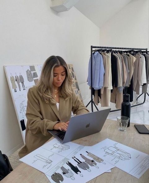 Designer working at laptop with fashion sketches and clothing rack, creating modern apparel in a stylish office.