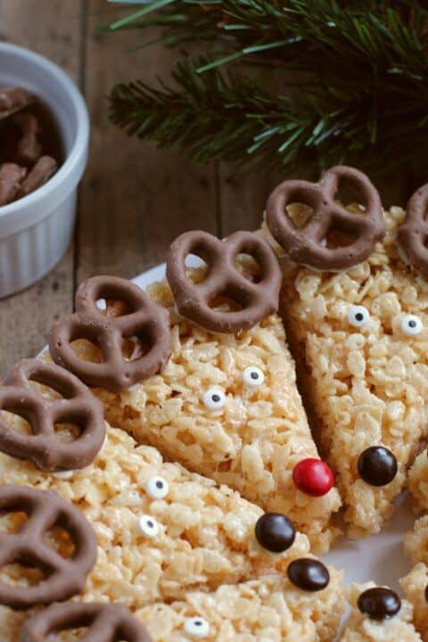 Reindeer-themed rice cereal treats with chocolate pretzel antlers and candy eyes on a festive plate.