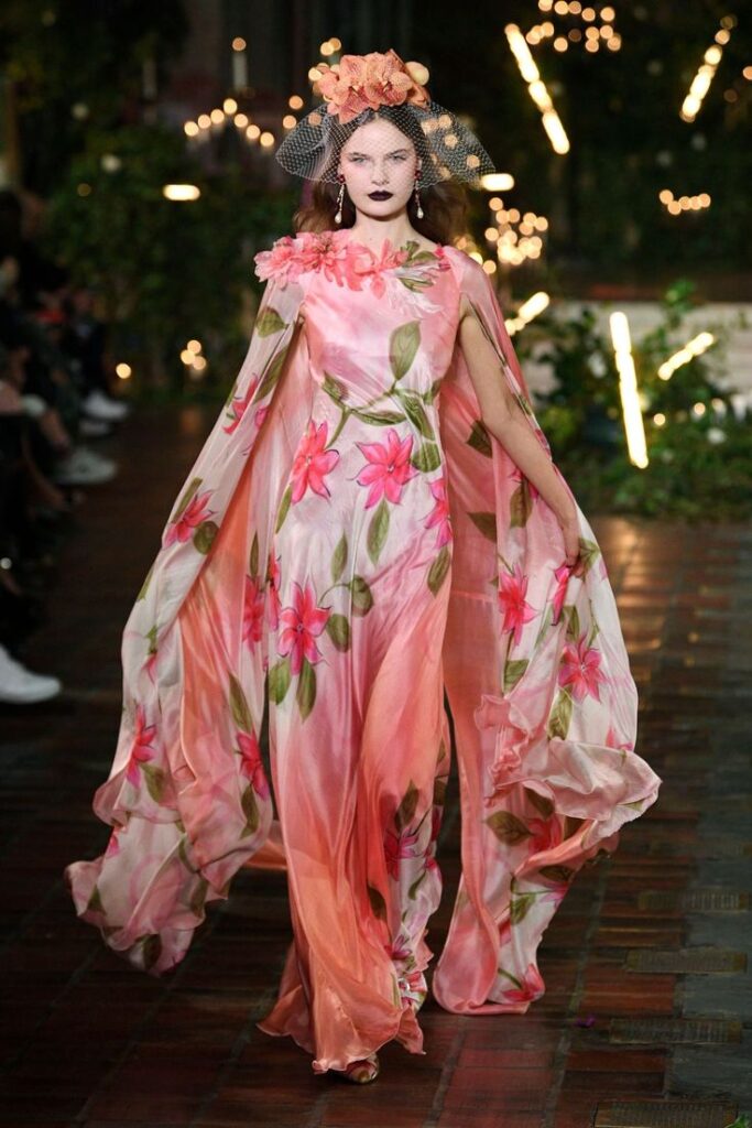 Rodarte Fall-Winter 2020 RTW Model in a flowing floral pink gown with headpiece walks runway, surrounded by dim lights and greenery. | Sky Rye Design Model in a flowing floral pink gown with headpiece walks runway, surrounded by dim lights and greenery.