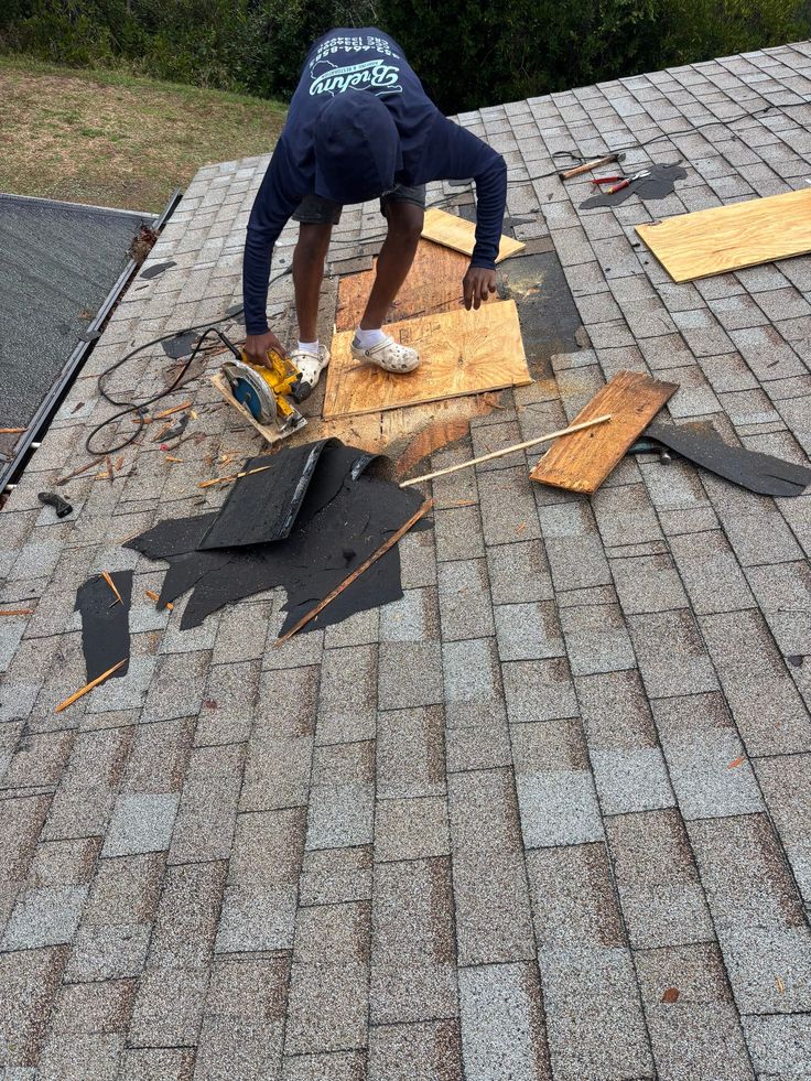 Worker repairing roof shingles with tools and plywood sheets, ensuring durable roof maintenance and safety.