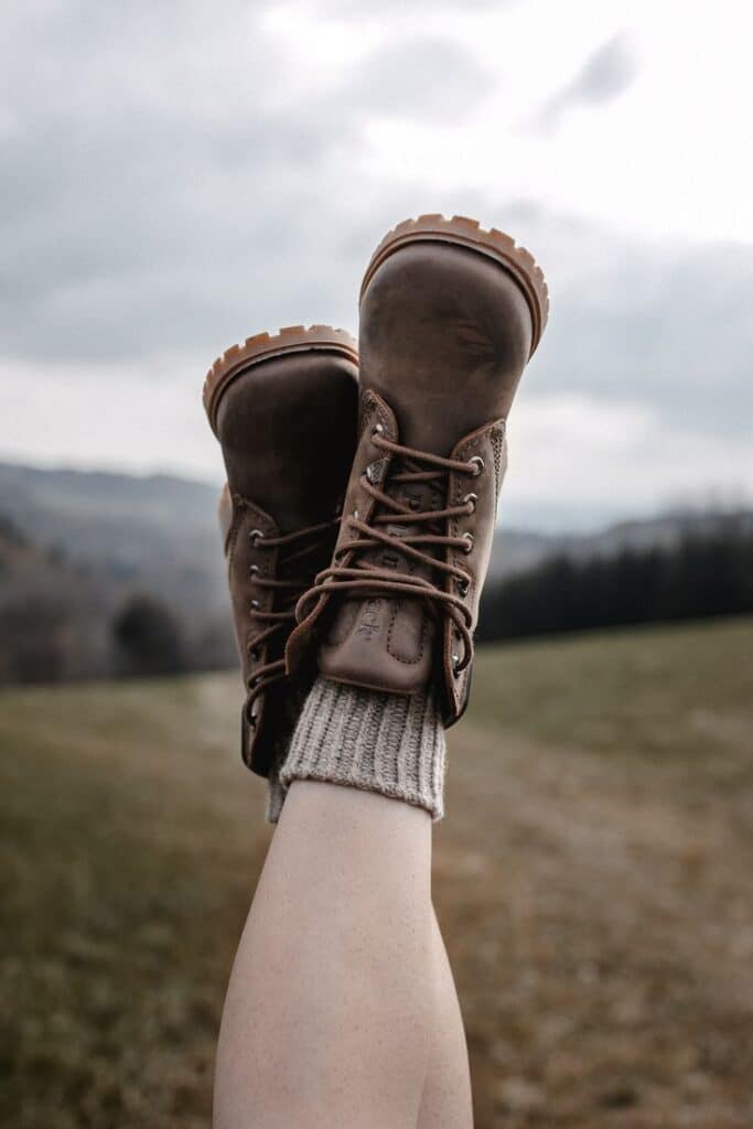 Legs raised in brown hiking boots and cozy socks against a cloudy sky in a grassy landscape. Outdoor adventure vibe.