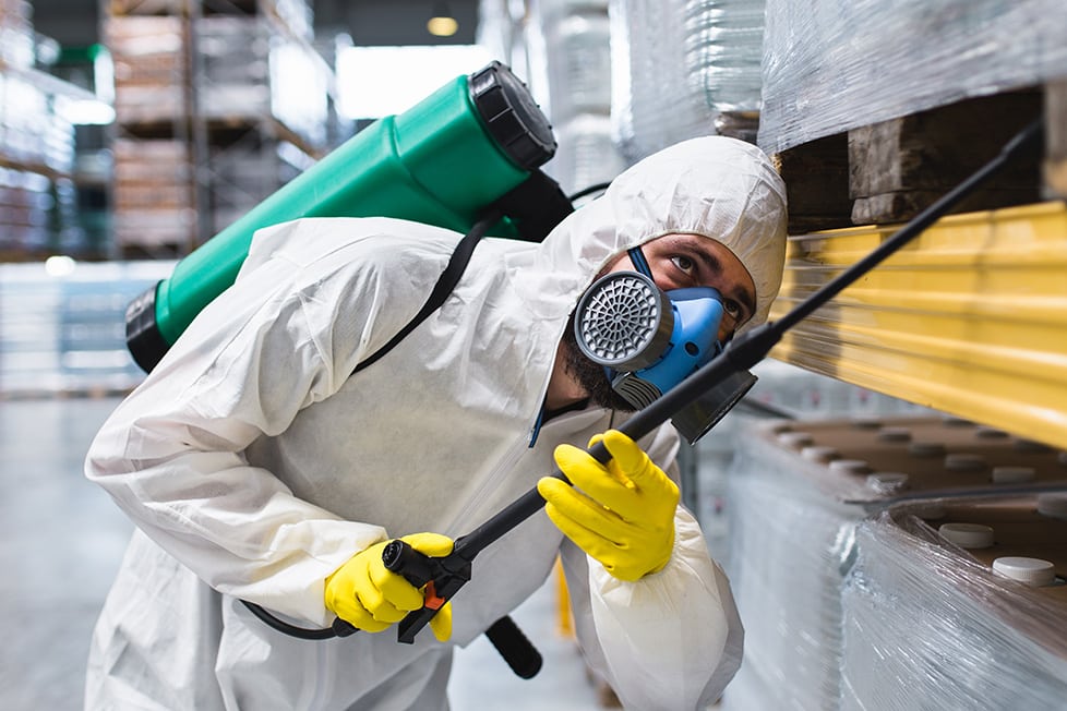 Pest control worker in protective gear spraying warehouse shelves, ensuring safety from infestations.