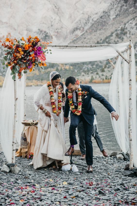 Outdoor wedding ceremony by a lake with couple under floral arch, wearing traditional garlands.