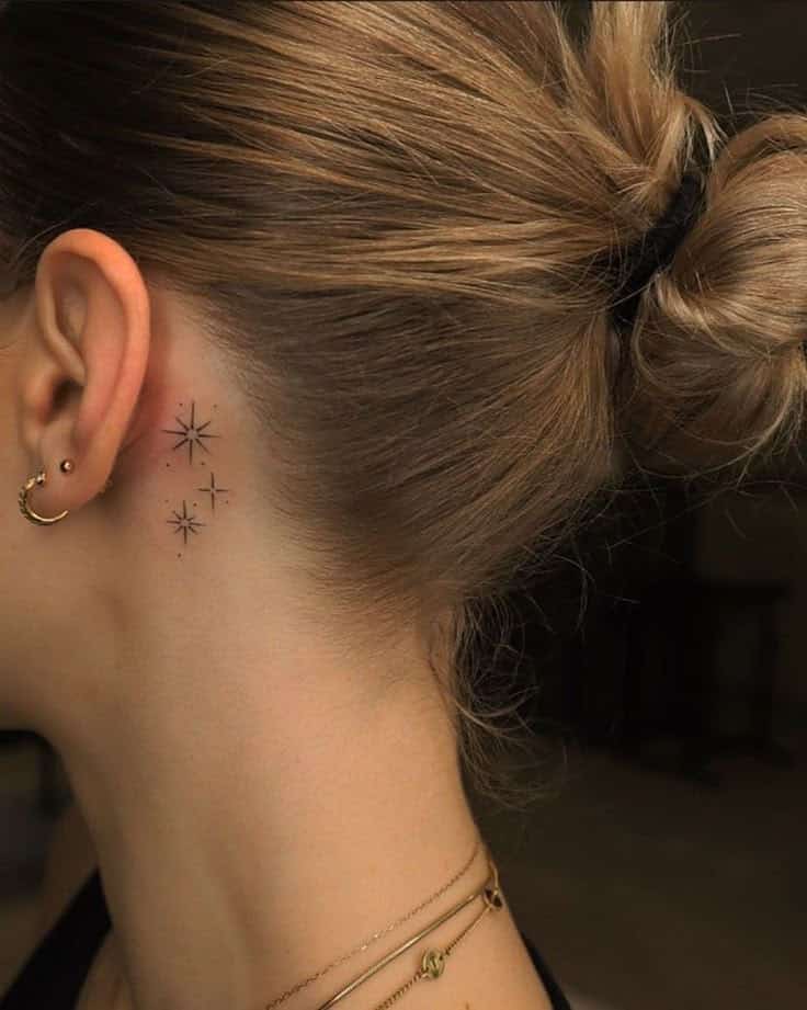 Star tattoos behind ear on woman with blond hair tied up, wearing gold earrings and necklaces.