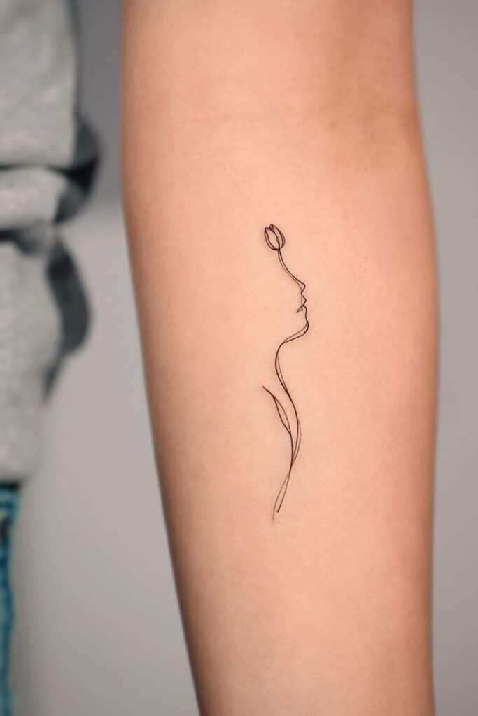 Minimalist tattoo of a flower integrated with a line drawing of a face on a person's forearm.