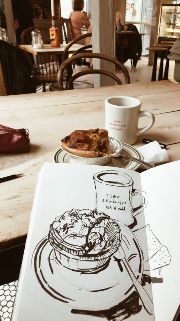 Sketchbook with pie illustration and coffee cup on cafe table, cozy atmosphere.