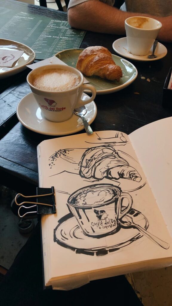 Sketch of a croissant and coffee beside real croissant and coffee on a café table.
