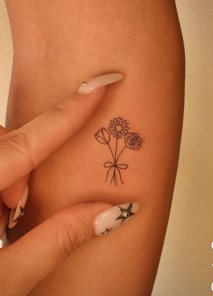 Minimalist flower tattoo on arm with daisies and roses, highlighted by star-adorned nails.