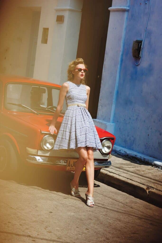 Woman in a vintage dress leaning on a classic red car in a sunny urban street scene.