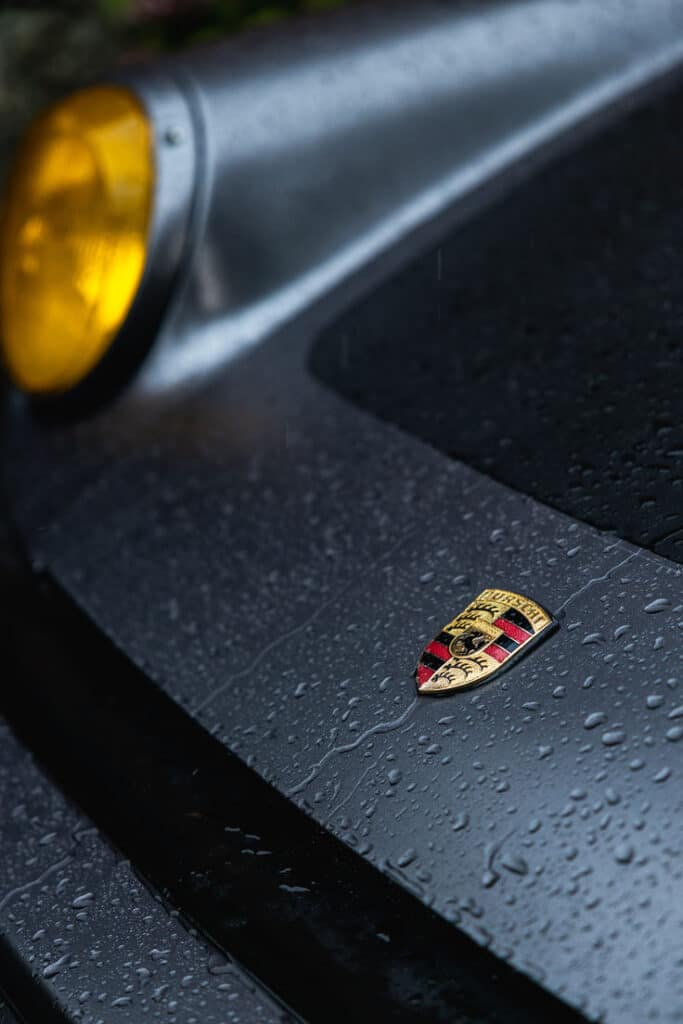 Close-up of a wet car hood with a Porsche emblem and yellow headlight, showcasing elegance and luxury.